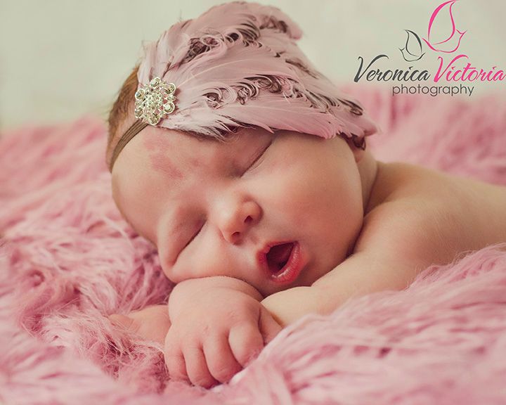 A sleeping baby wearing a pink feathered headband lies on soft pink fabric during a newborn photo session.