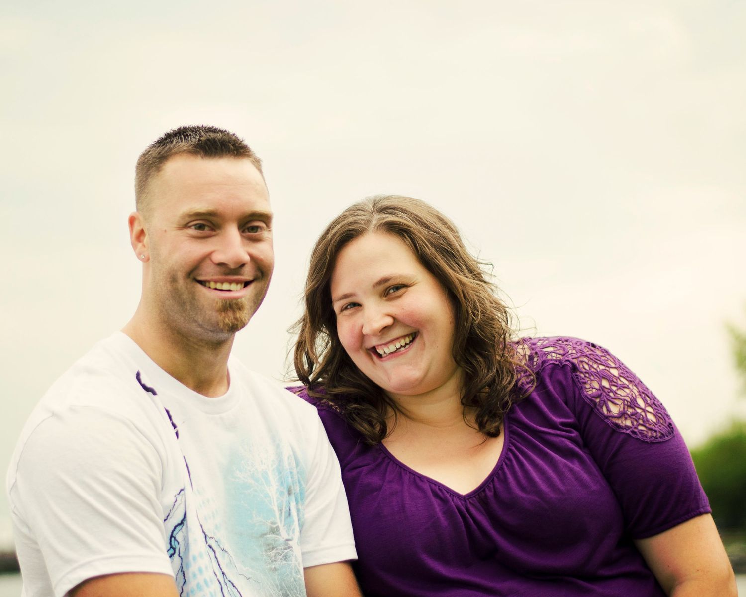 A couple in white and purple outfits smiling and embracing while posing for outdoor engagement photos.