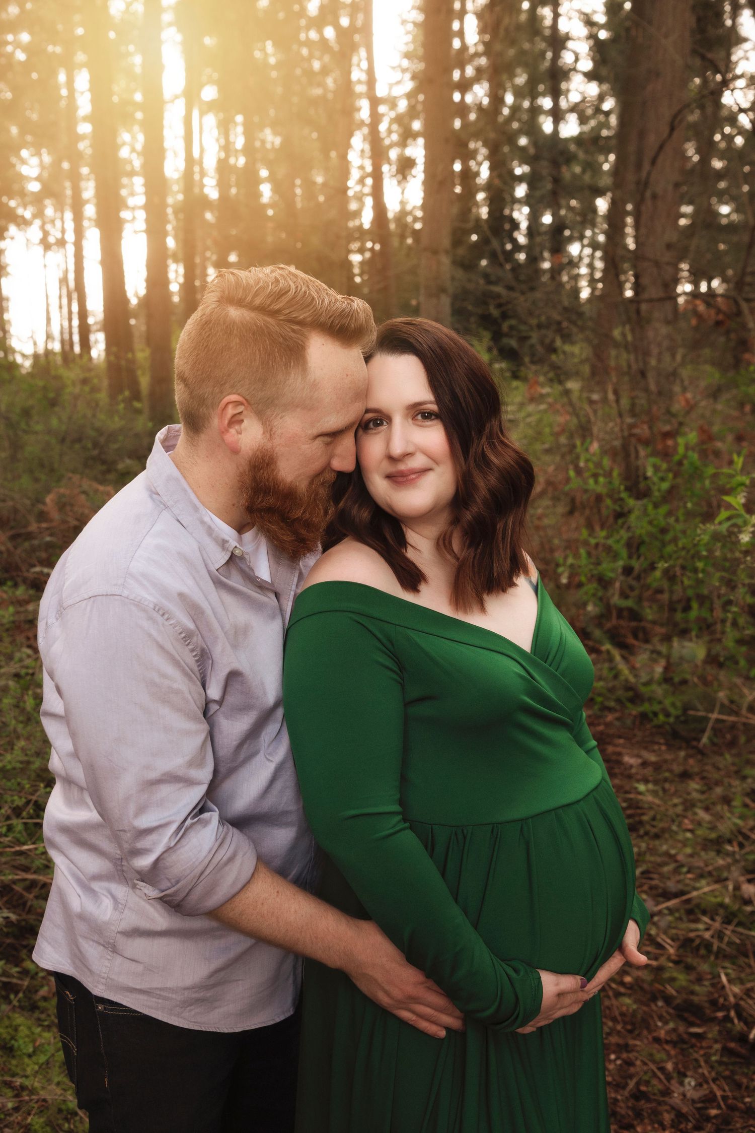 A maternity photo of a couple in a forest with golden sunlight streaming through the trees.