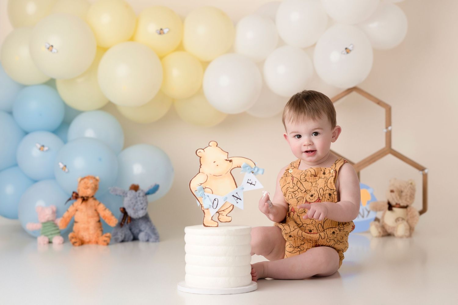 Baby in yellow outfit sits near first birthday cake with blue and yellow balloon decorations.