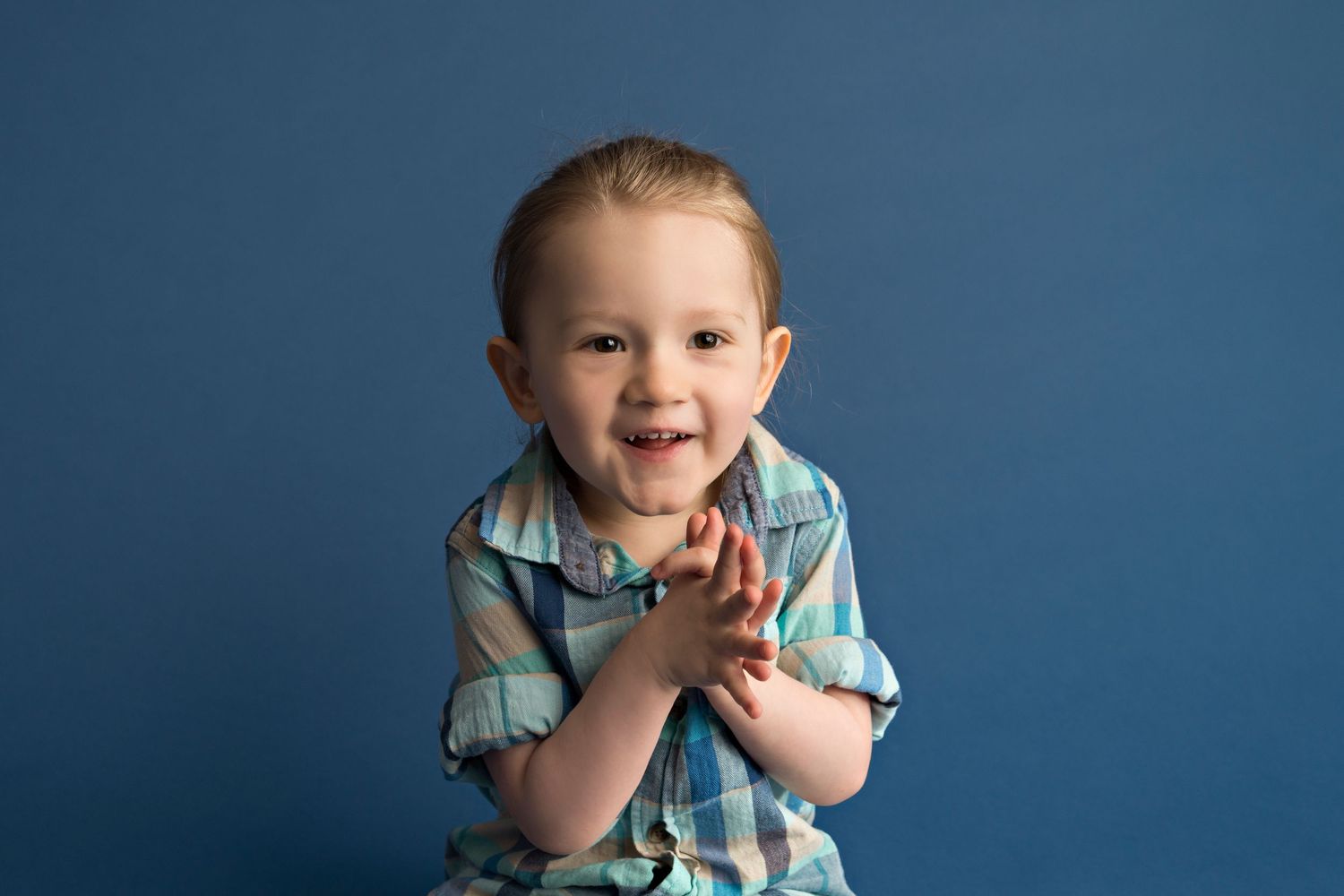 Young child in a plaid shirt clapping and smiling against a blue studio background.