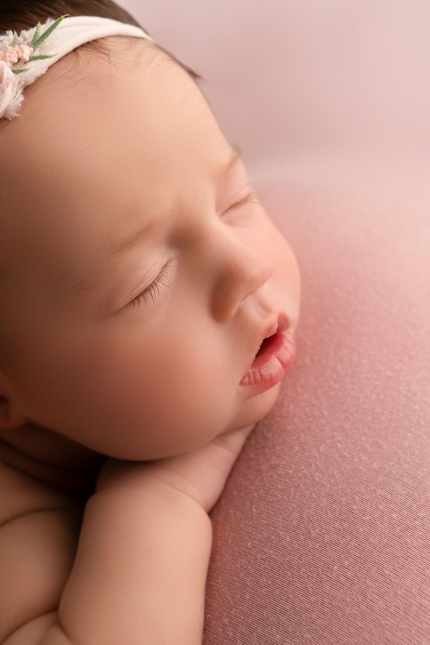 A peaceful sleeping newborn rests on soft pink fabric wearing a delicate white headband.