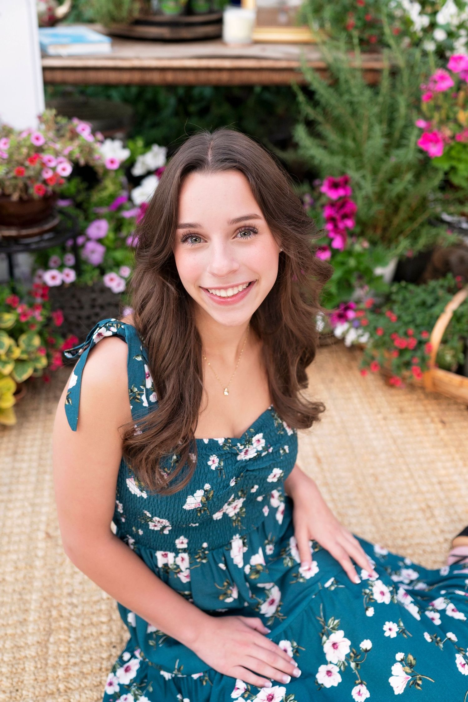 Woman in teal floral dress sitting outdoors surrounded by colorful potted flowers and greenery.
