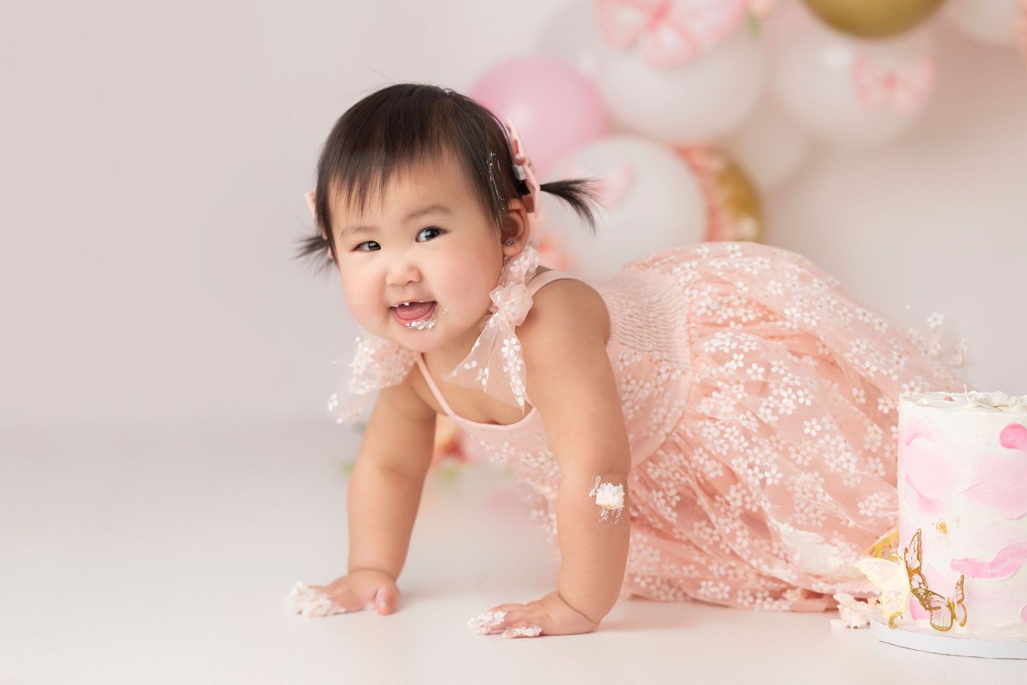 Baby in pink lace dress crawls on white floor with pink balloon decorations.
