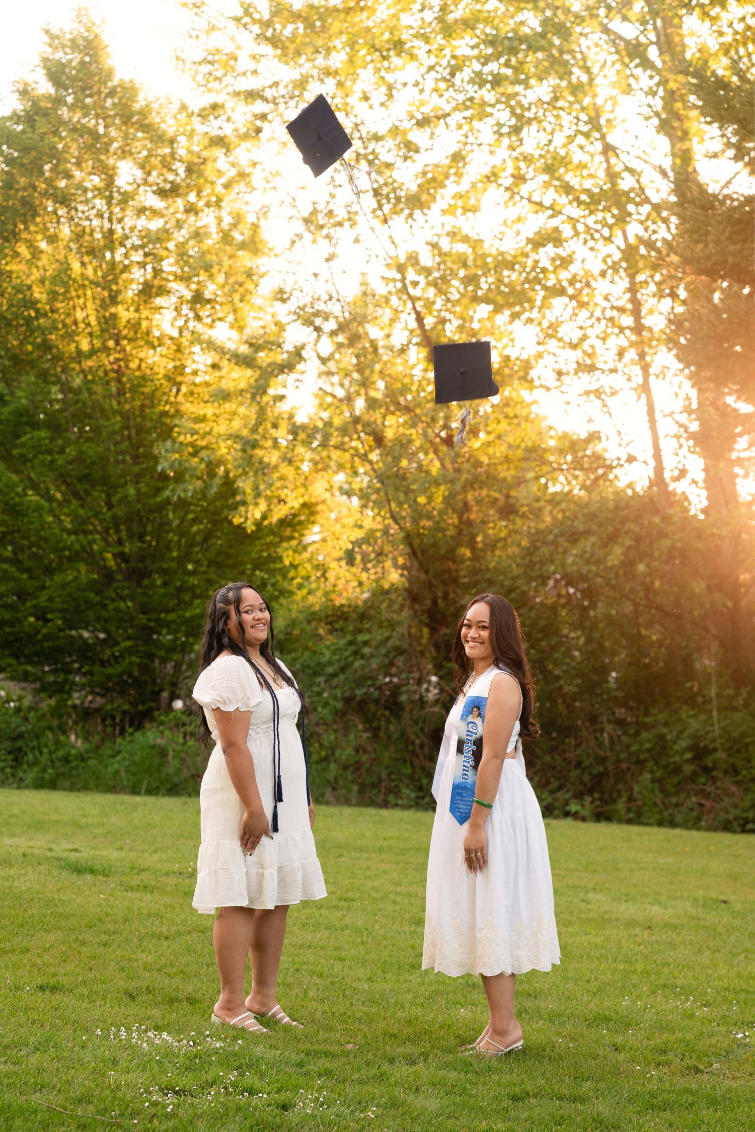 Two graduates in white dresses toss their caps in the air on a sunny lawn.