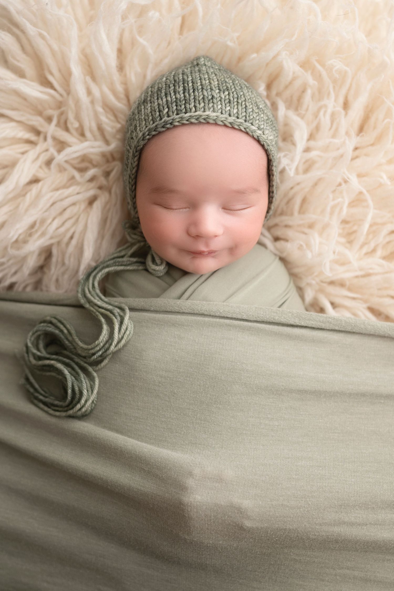 Sleeping newborn swaddled in soft sage green fabric wearing a matching knit bonnet on cream fur backdrop.