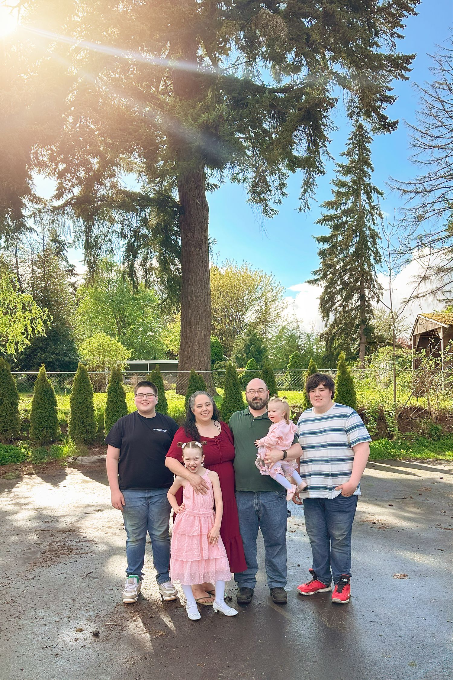 Outdoor family photo in park with tall evergreen trees and sunlight.