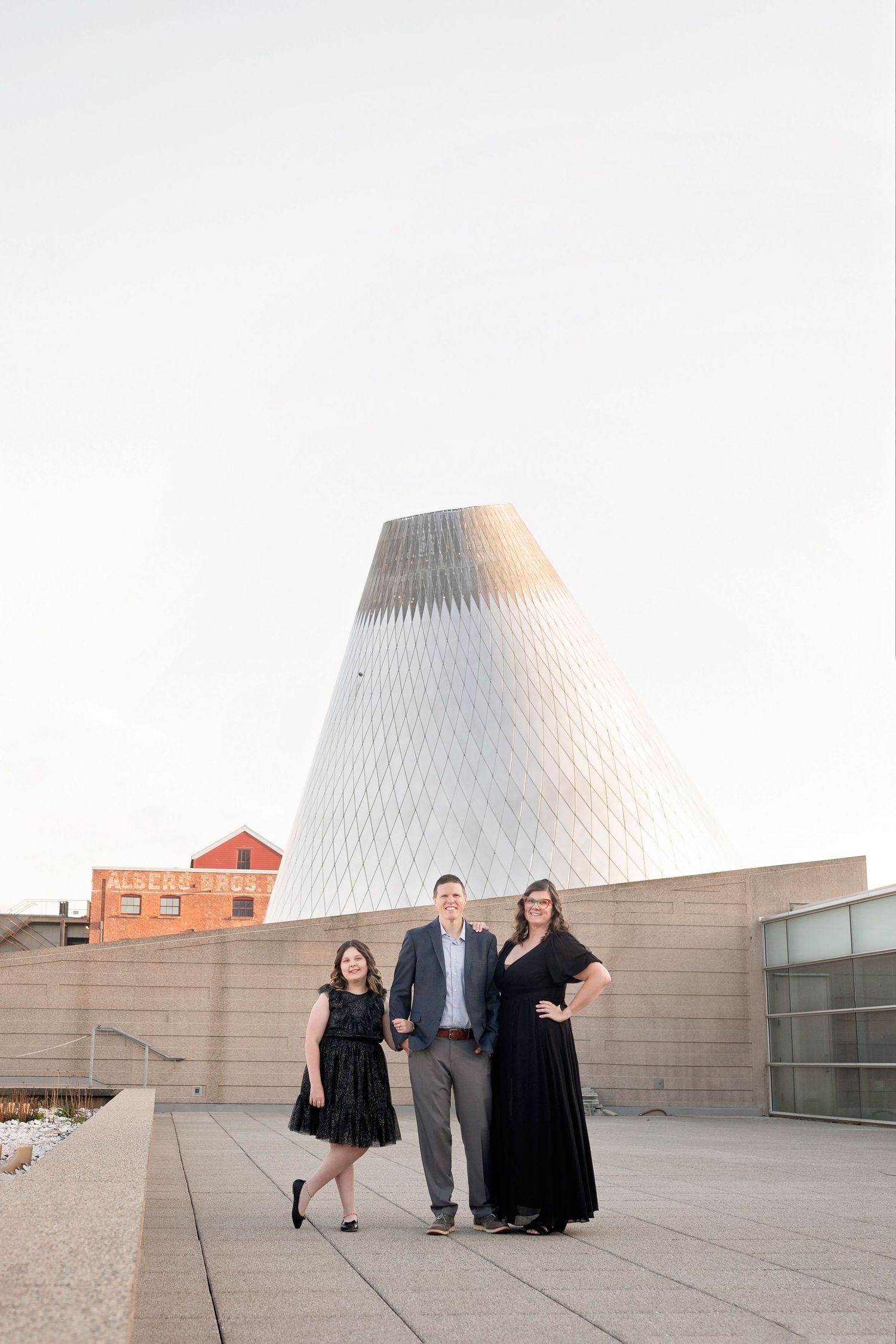 Three people posing in formal attire outside modern architecture building with cone-shaped structure.