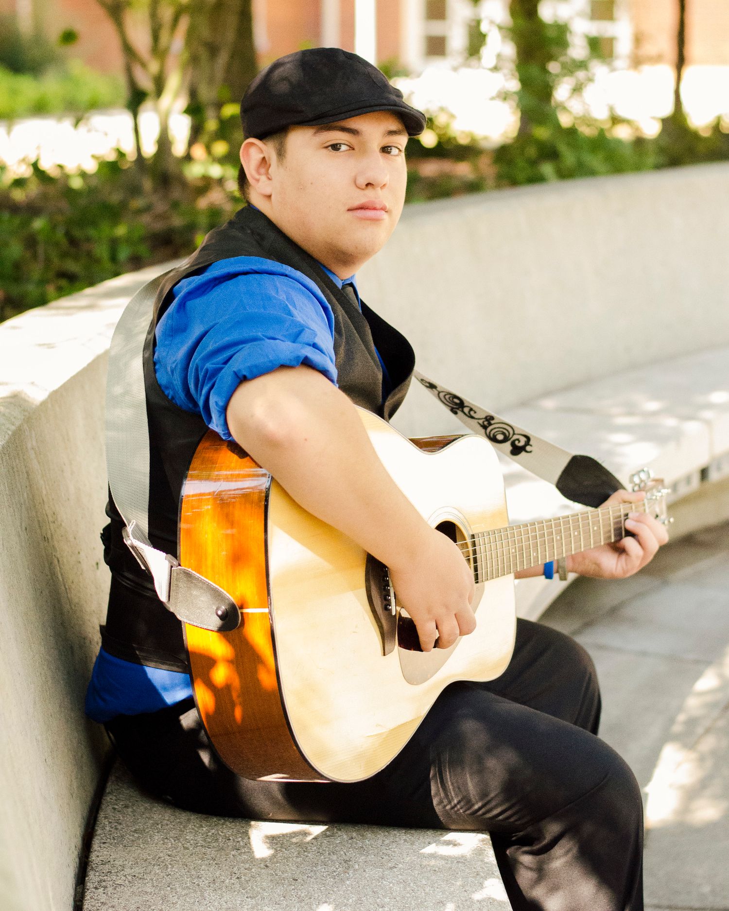 Musician in a black cap and blue shirt plays acoustic guitar while sitting outdoors on stone steps.