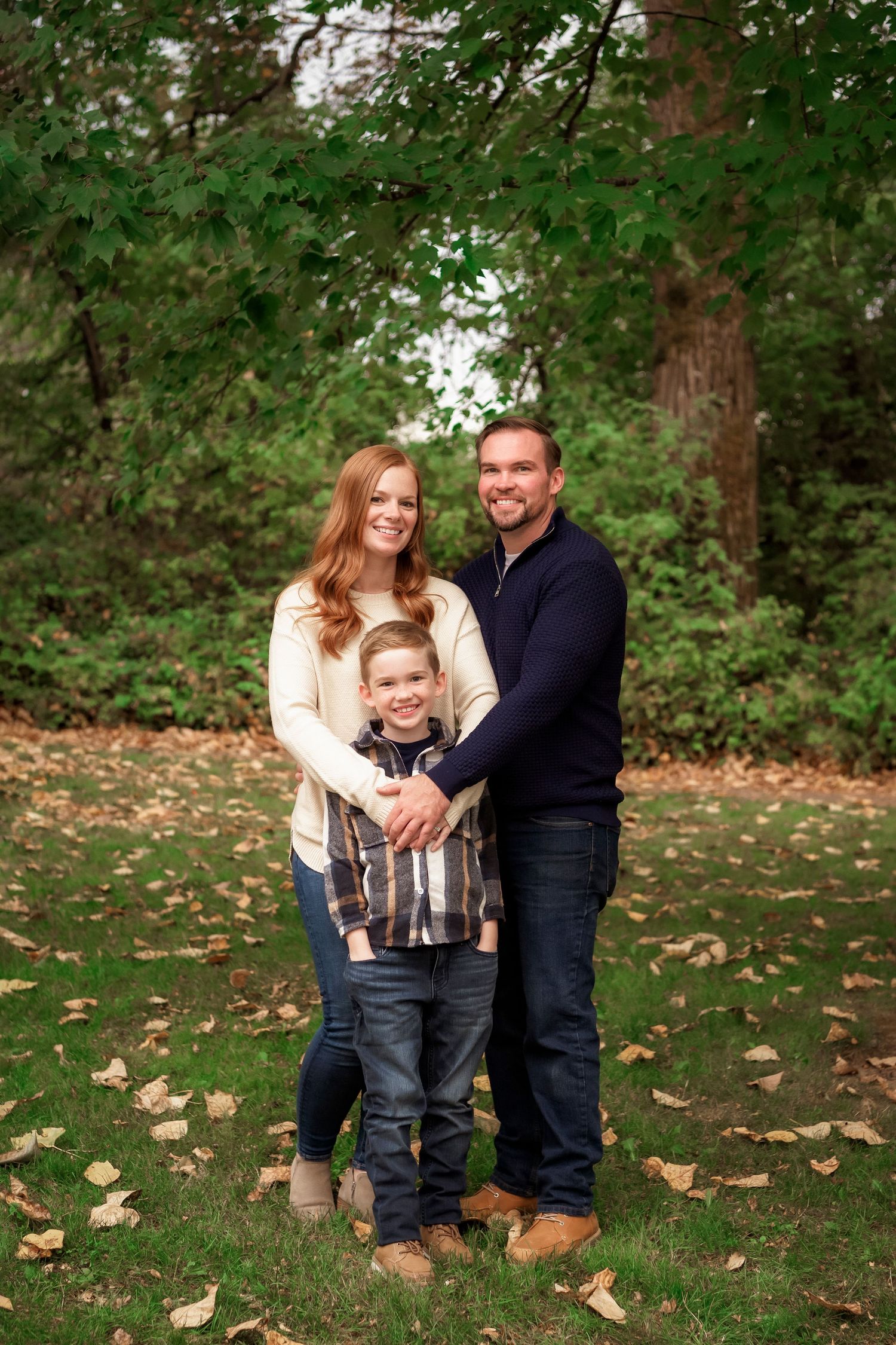 A family of three poses together on fallen autumn leaves in a wooded setting.
