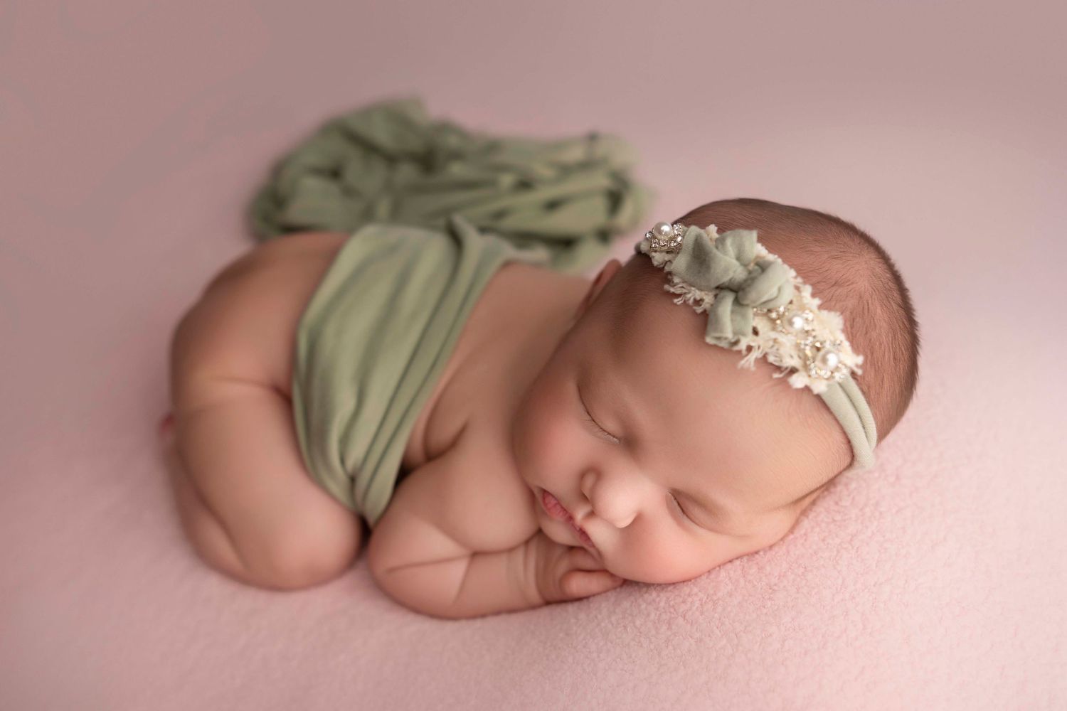 Newborn baby sleeping peacefully on pink backdrop wearing sage green wrap and delicate floral headband.