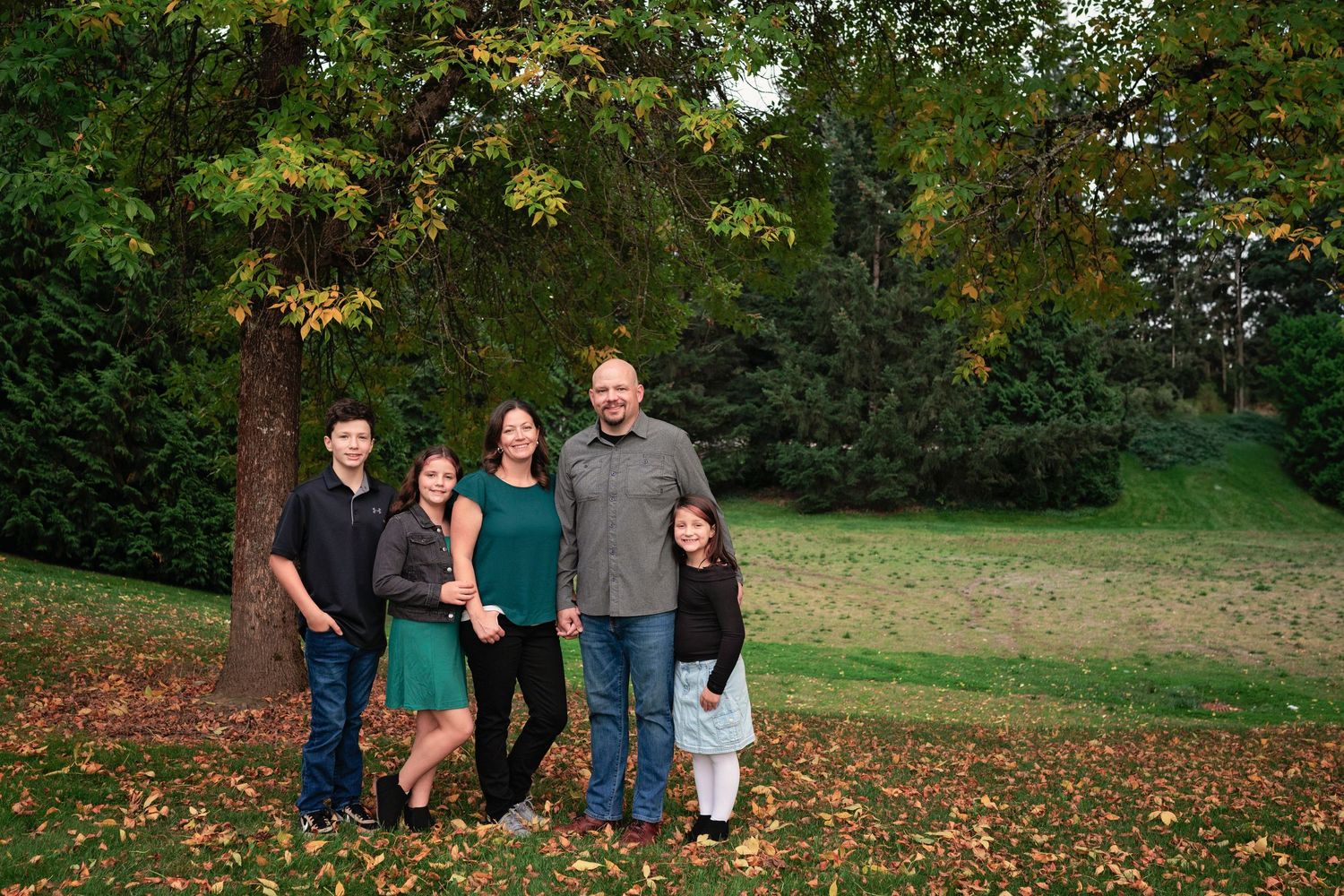 A family poses together for a portrait on fallen autumn leaves with trees in background.