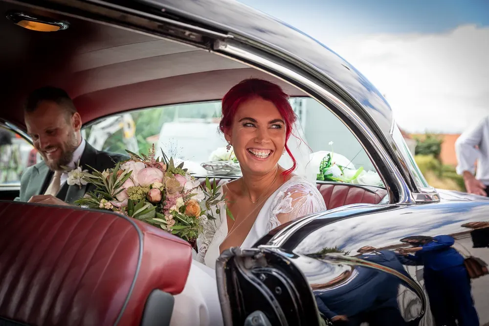 Bride with bright red hair sits in vintage car with red leather interior.