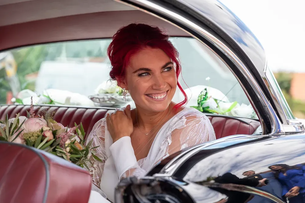 Happy bride with red hair poses inside classic car with red leather seats.