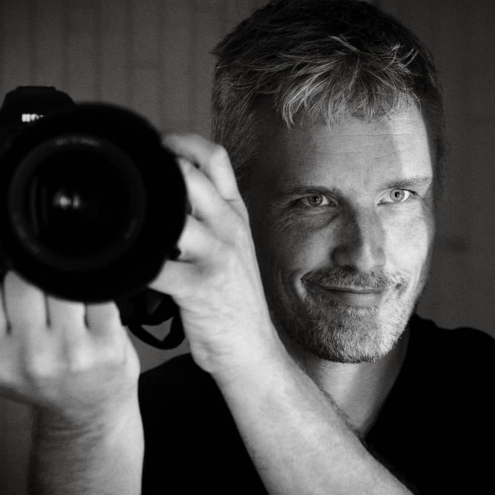 Black and white portrait of a photographer holding a professional camera with a telephoto lens.