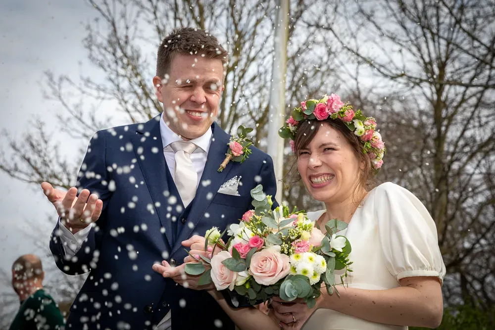 A couple celebrates their wedding day with confetti in front of bare winter trees.