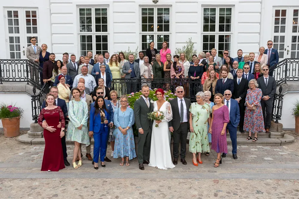 Large wedding party group photo on the steps of an elegant white building with ornate windows and railings.