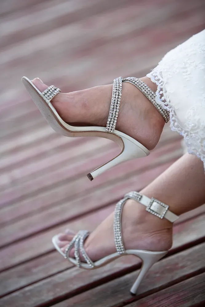 Elegant rhinestone-studded white wedding heels on wooden deck.