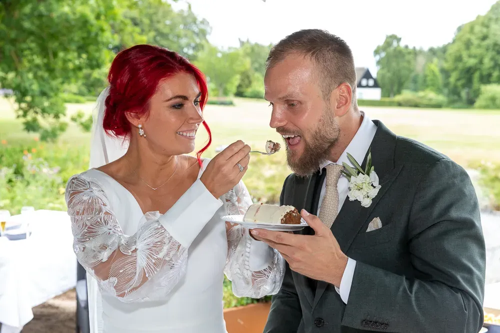 Couple sharing cake at an outdoor wedding reception with green scenery in background.
