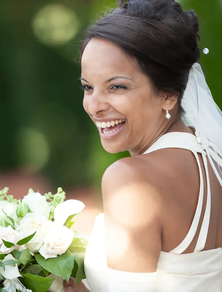 A joyful bride laughs while holding white flowers against a green background.