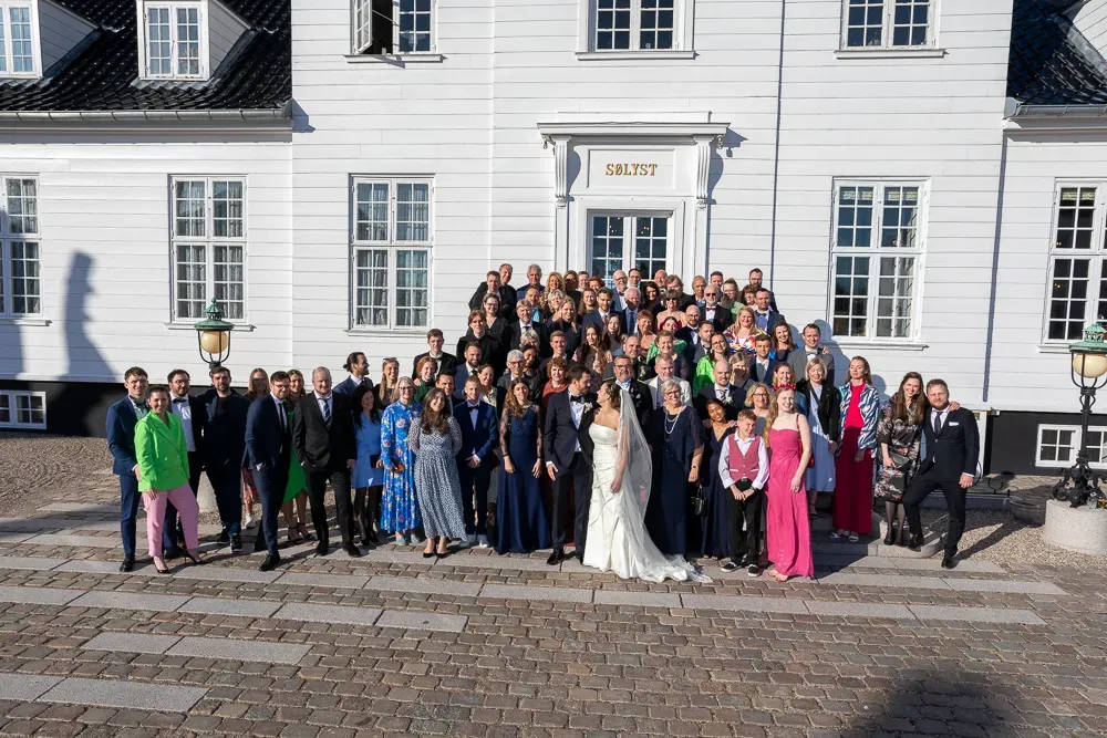 Large wedding group photo in front of a white mansion building with multiple windows.