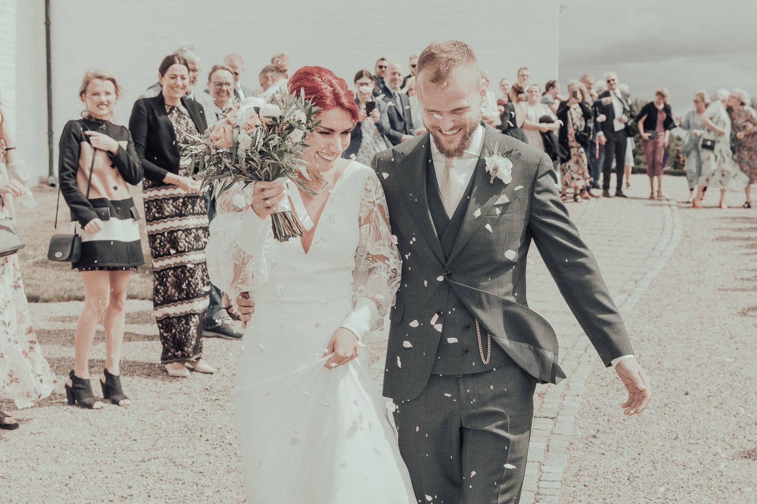 Happy newlyweds walk through shower of confetti after outdoor wedding ceremony as guests celebrate.
