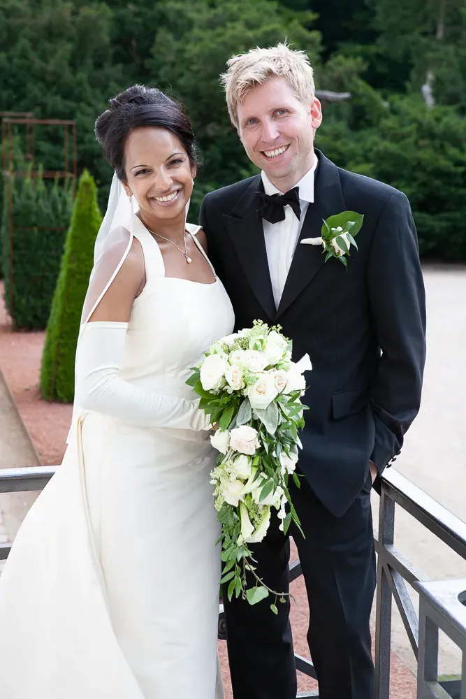 A bride in white dress holds cascading bouquet next to groom in tuxedo on garden steps.