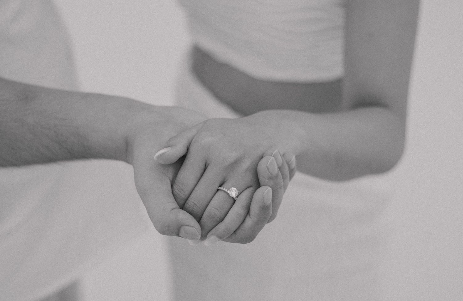 Close-up black and white image of two hands clasped together, with a focus on the hand wearing an engagement ring. The r
