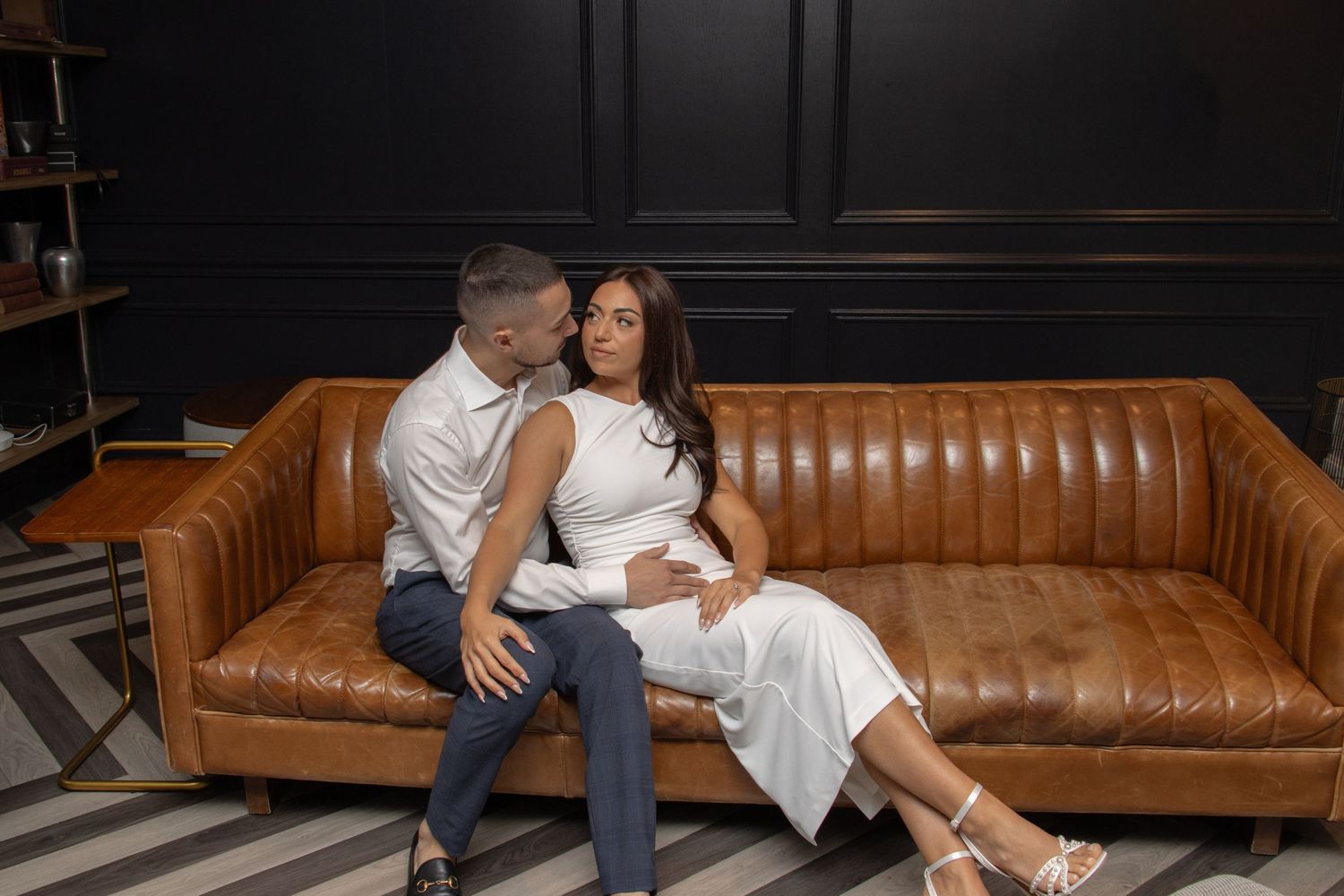 A couple sits on a modern sofa in an elegant, dimly lit room. The man, in a dress shirt and pants, gently holds the woma