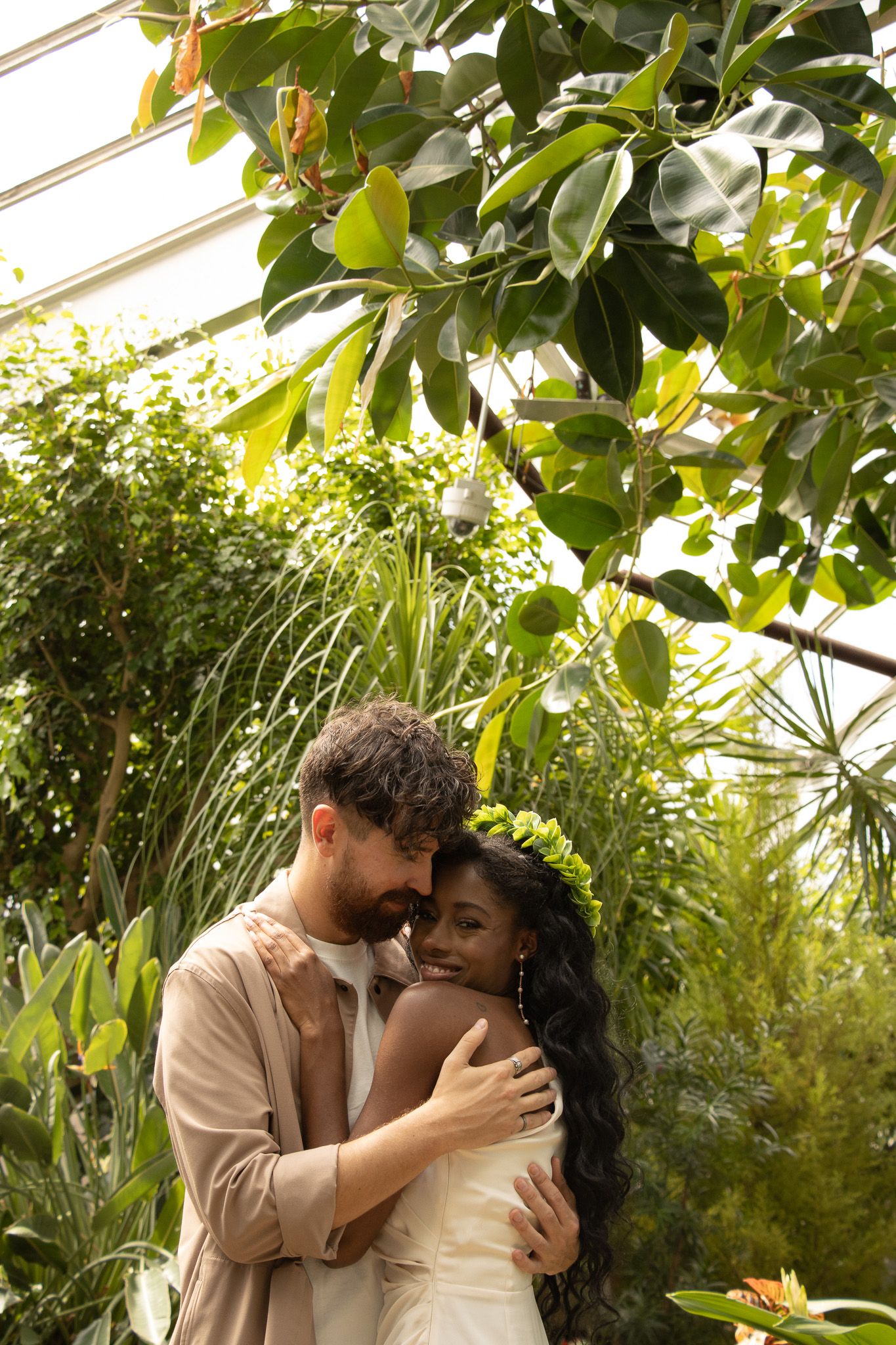 A couple embraces in a lush garden. The man, with curly hair and a beard, smiles while holding the woman, who has long,