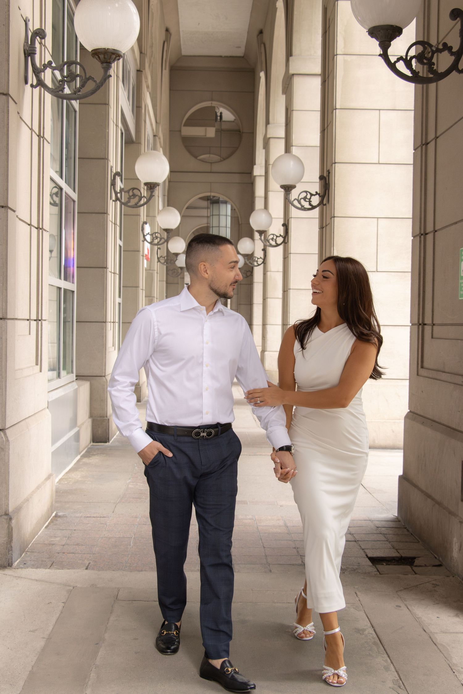 A couple walks hand in hand through a corridor lined with decorative lamps and arched windows. The man wears a white shi