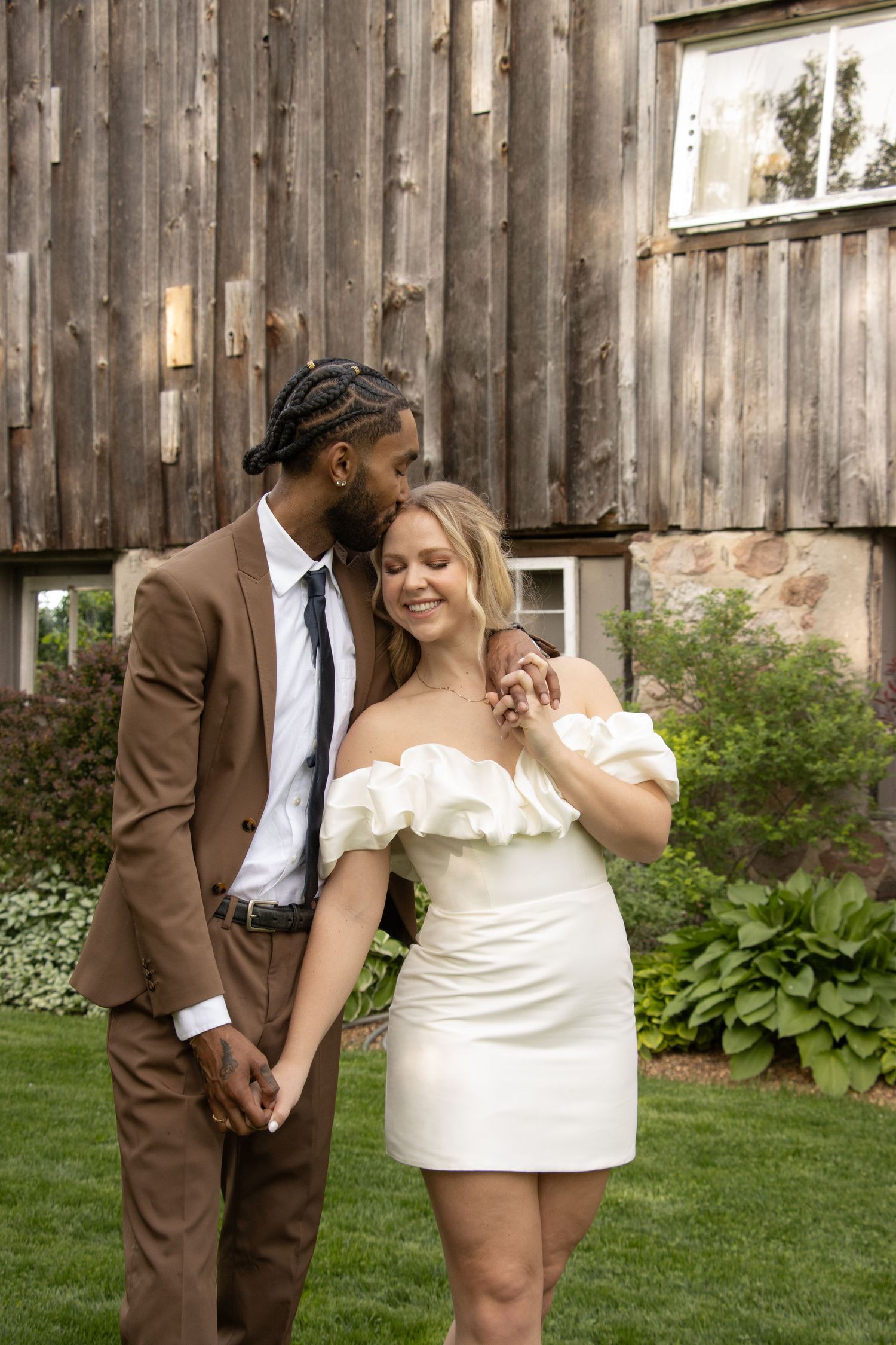 A man in a brown suit kisses the forehead of a woman in a short off-shoulder white dress. They are holding hands and smi