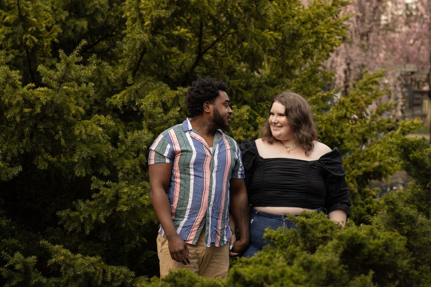 A woman in a black off-the-shoulder top and jeans holds hands with a man in a striped shirt and shorts. They are smiling