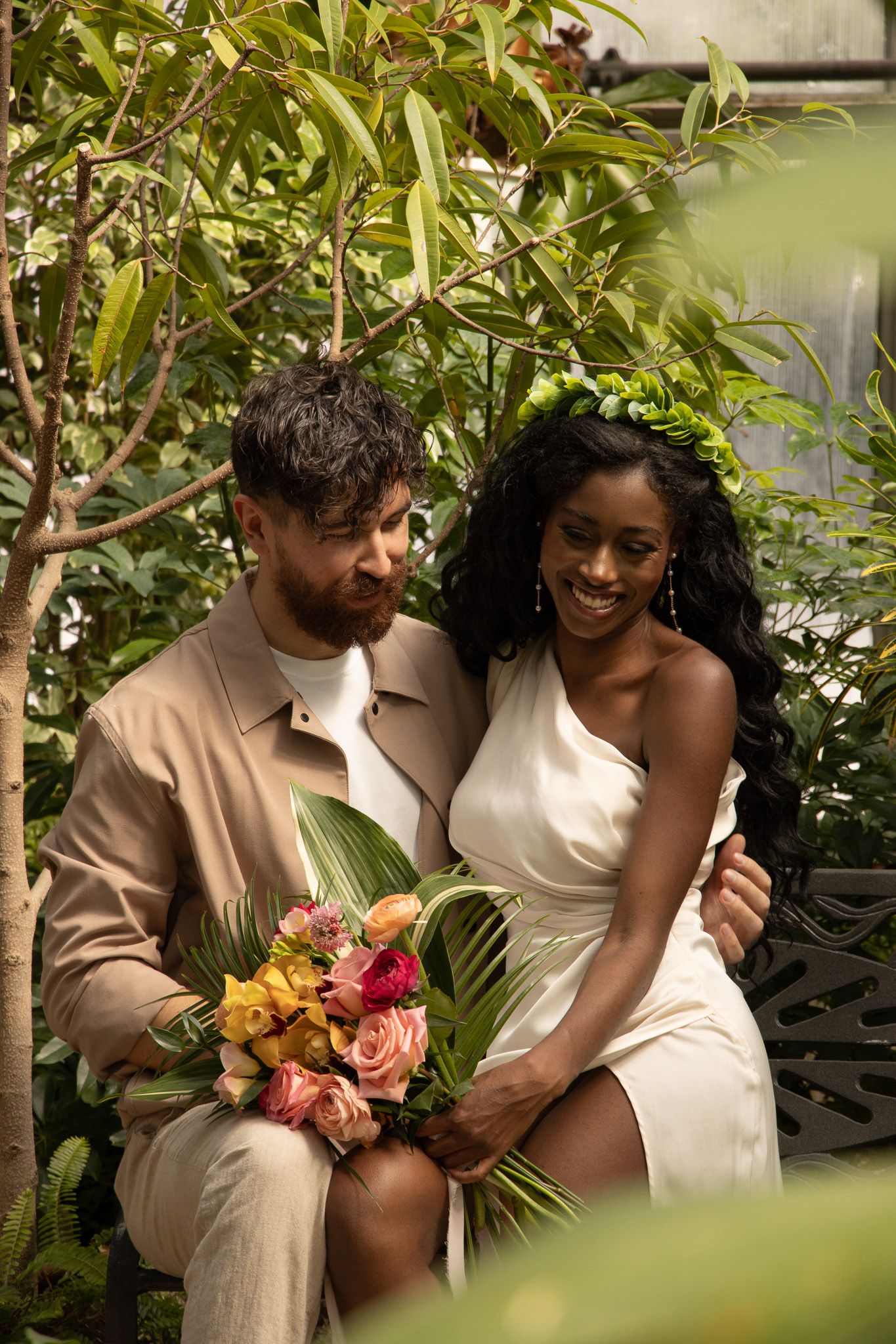 A man with a beard and a woman in a white dress are seated on a bench surrounded by greenery. They are smiling at each o