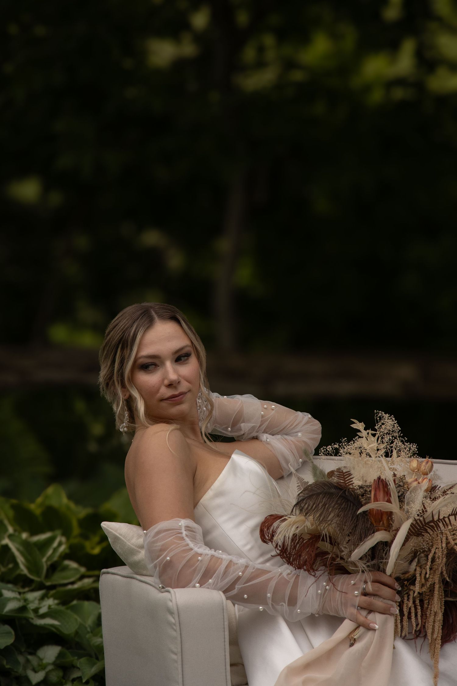 A bride in a strapless white wedding dress and sheer gloves sits on a white couch outdoors. She holds a bouquet featurin