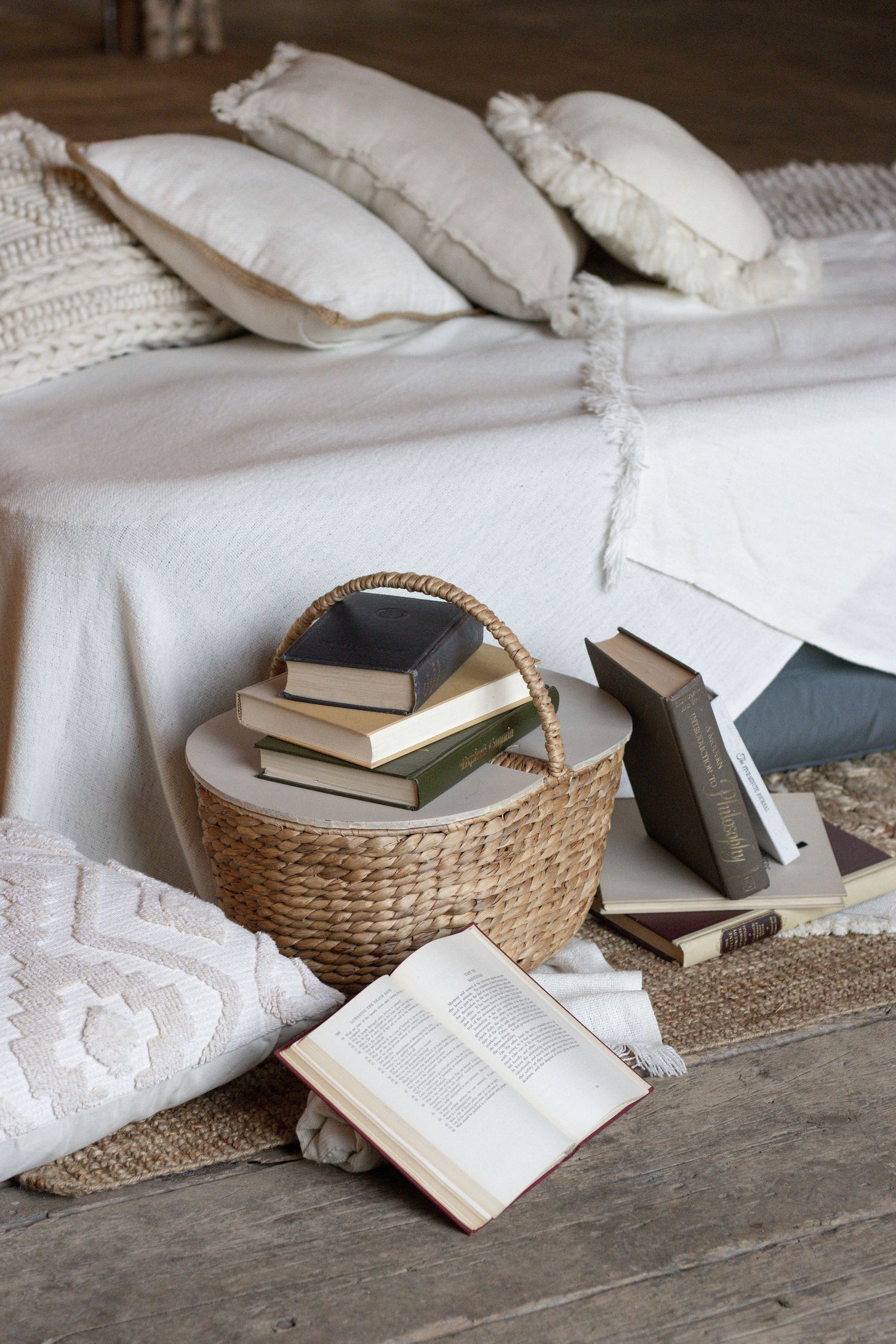A stack of vintage books on top of a wicker picnic basket. There are white texture blankets and pillows around.