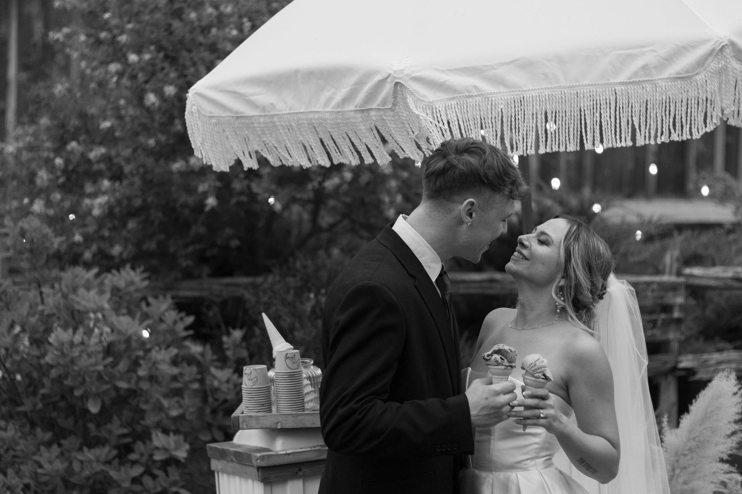 A black and white photo of a bride and groom. They are standing close, smiling at each other under a fringed umbrella. B