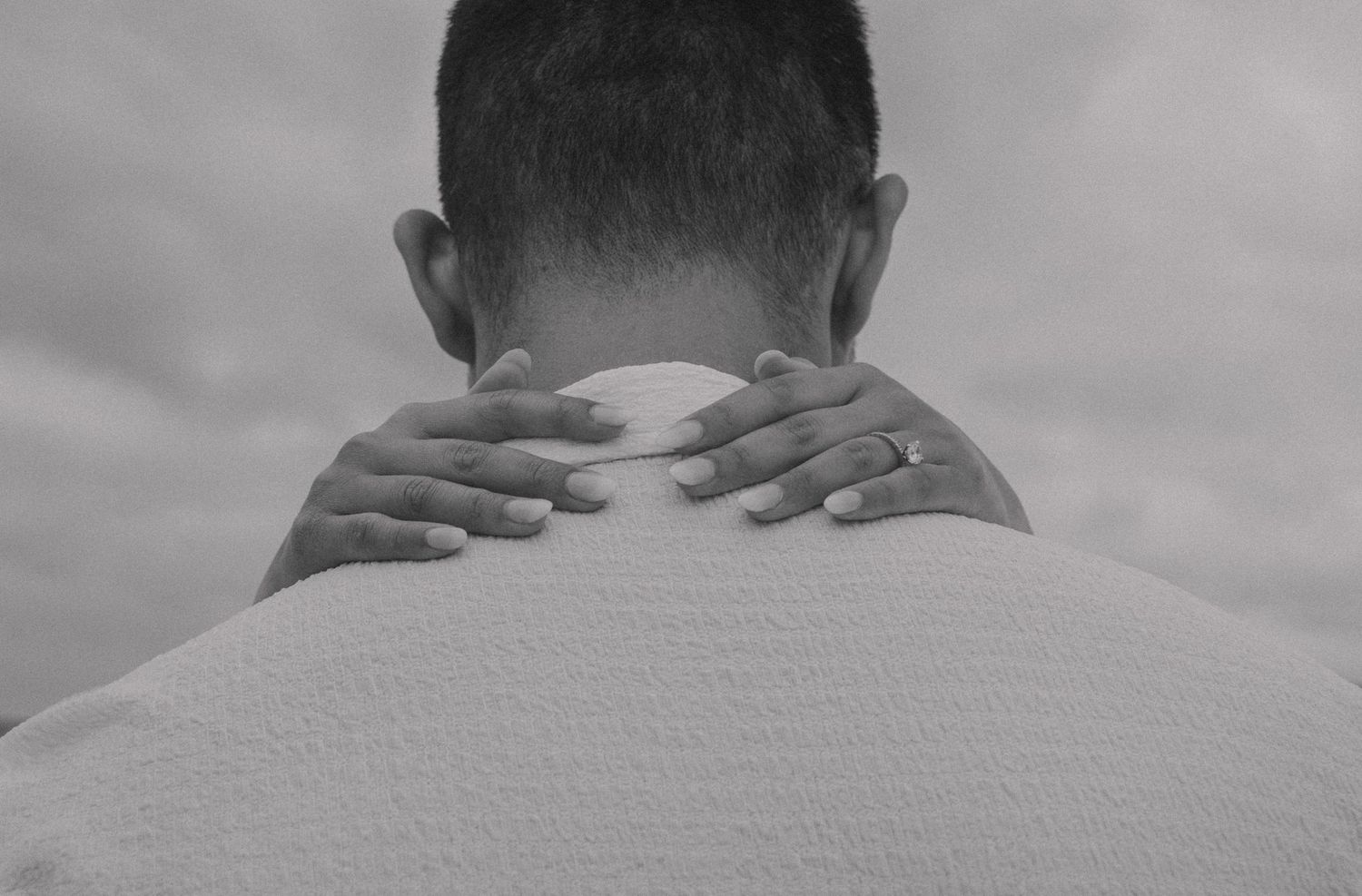 A close-up, black-and-white photo of a person from behind, with the focus on their neck and shoulders. Another person’s
