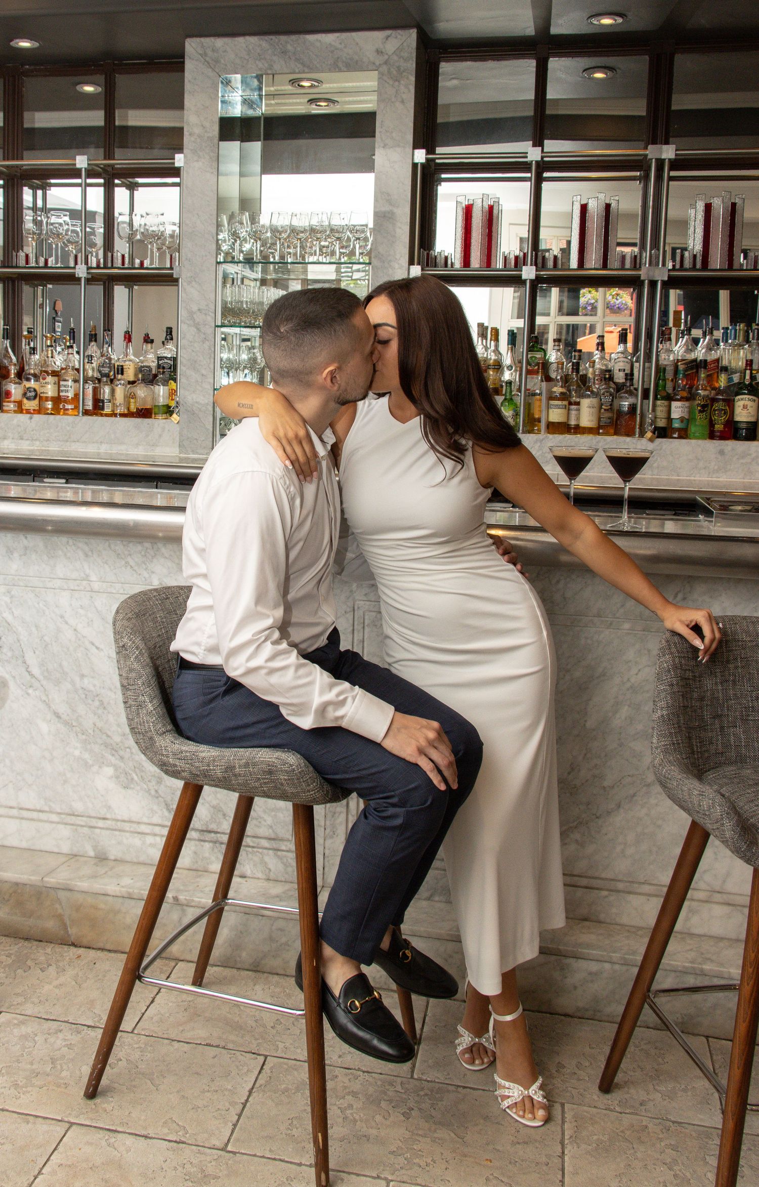 A man and a woman share a kiss while sitting at a bar. The man has short hair and wears a white shirt and dark pants. Th