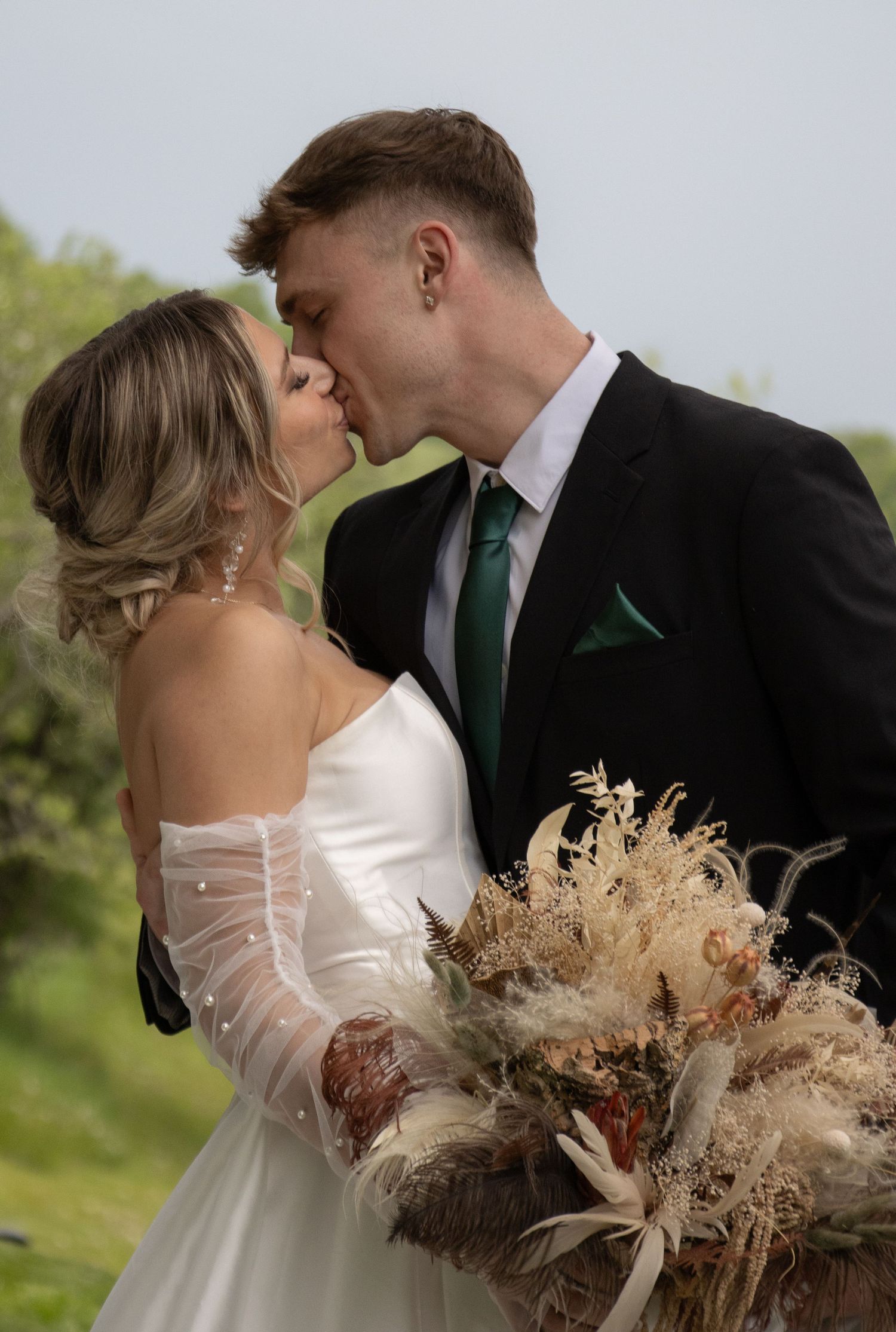 A couple dressed in wedding attire shares a kiss outdoors. The bride wears a white dress with off-the-shoulder sleeves a