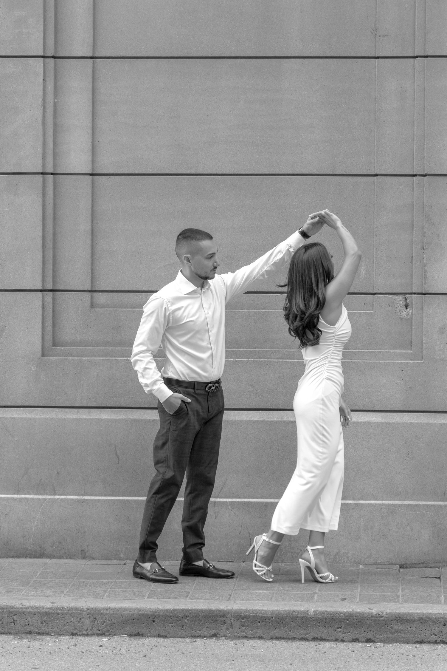 A man wearing a white shirt and dark pants is dancing with a woman in a white dress and high heels. He holds her hand an