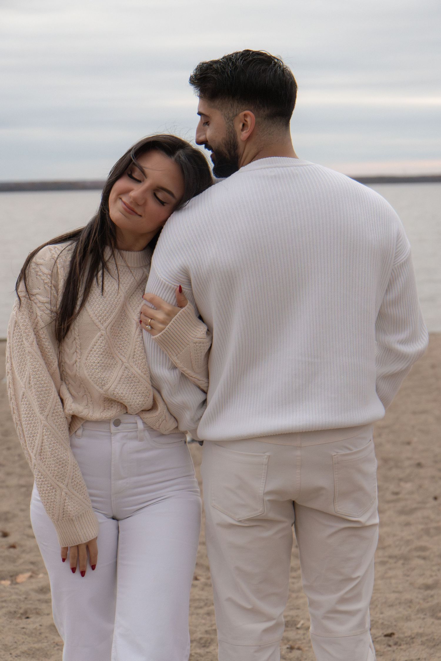 A man facing the water. A woman is facing forward with her arm around his arm. She is leaning her head on his shoulder.