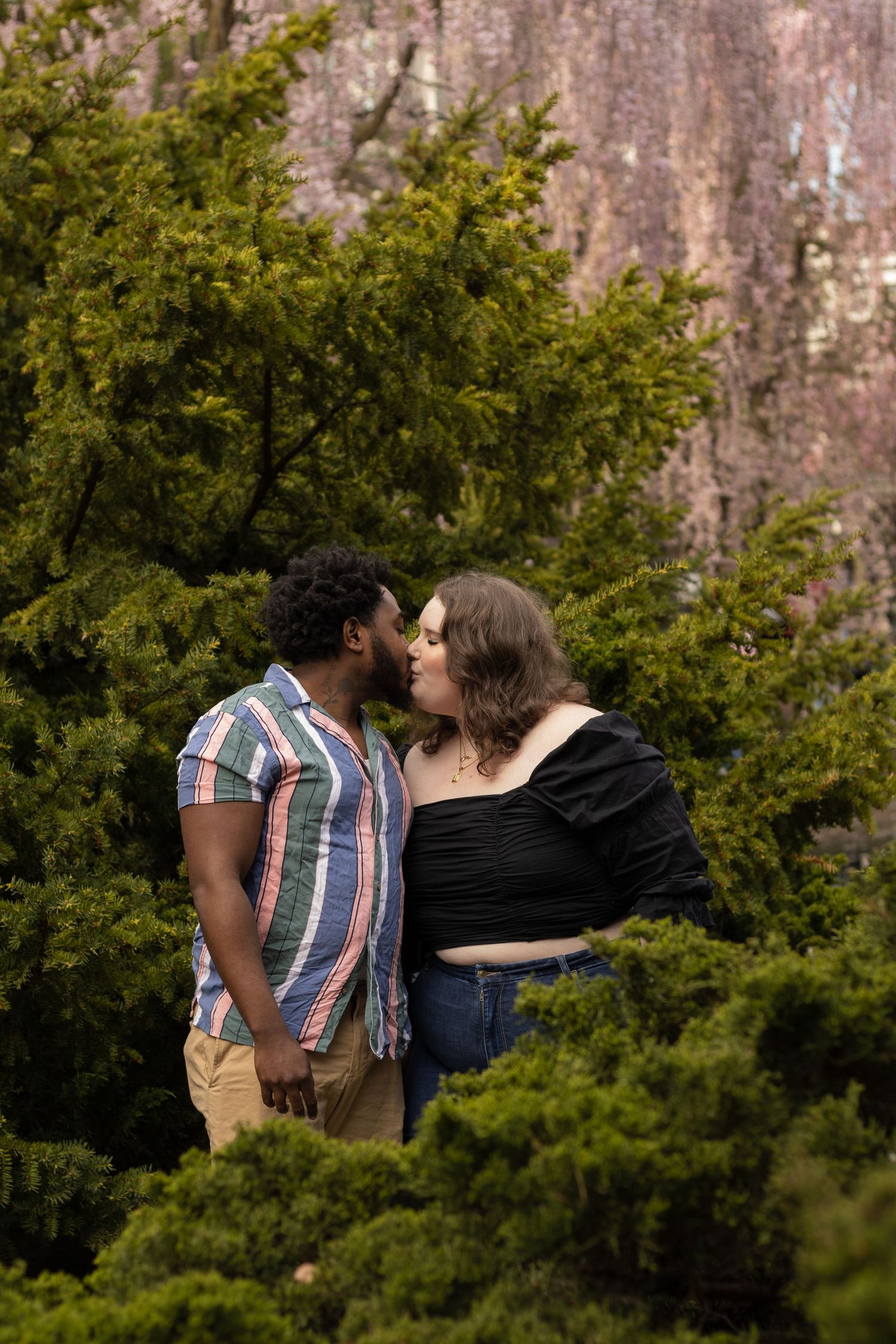 A woman in a black off-the-shoulder top and jeans holds hands with a man in a striped shirt and shorts. They are embraci