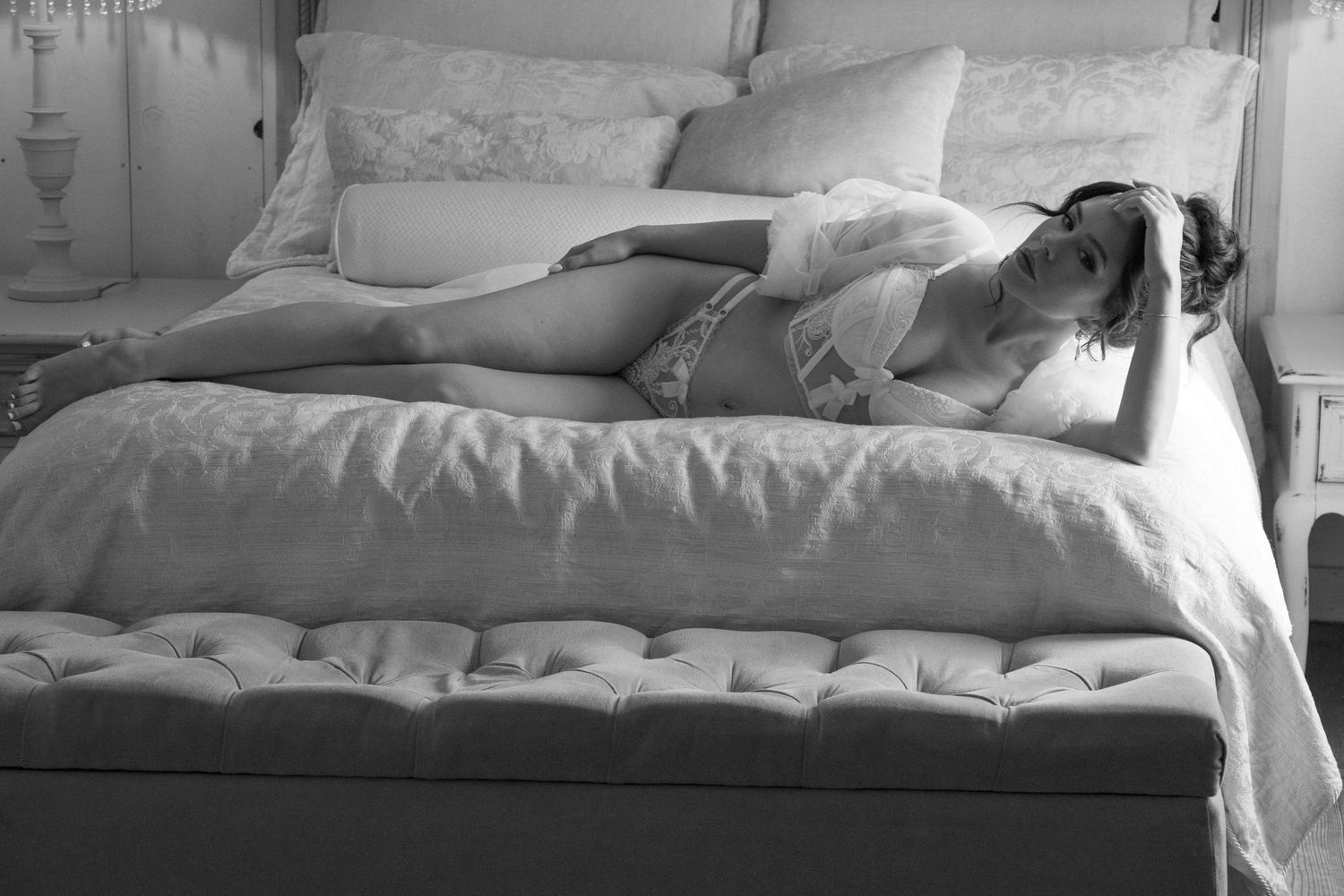 A woman in elegant lingerie reclines on a bed in a softly lit room. She is wearing a sheer top over a lacy bra and match