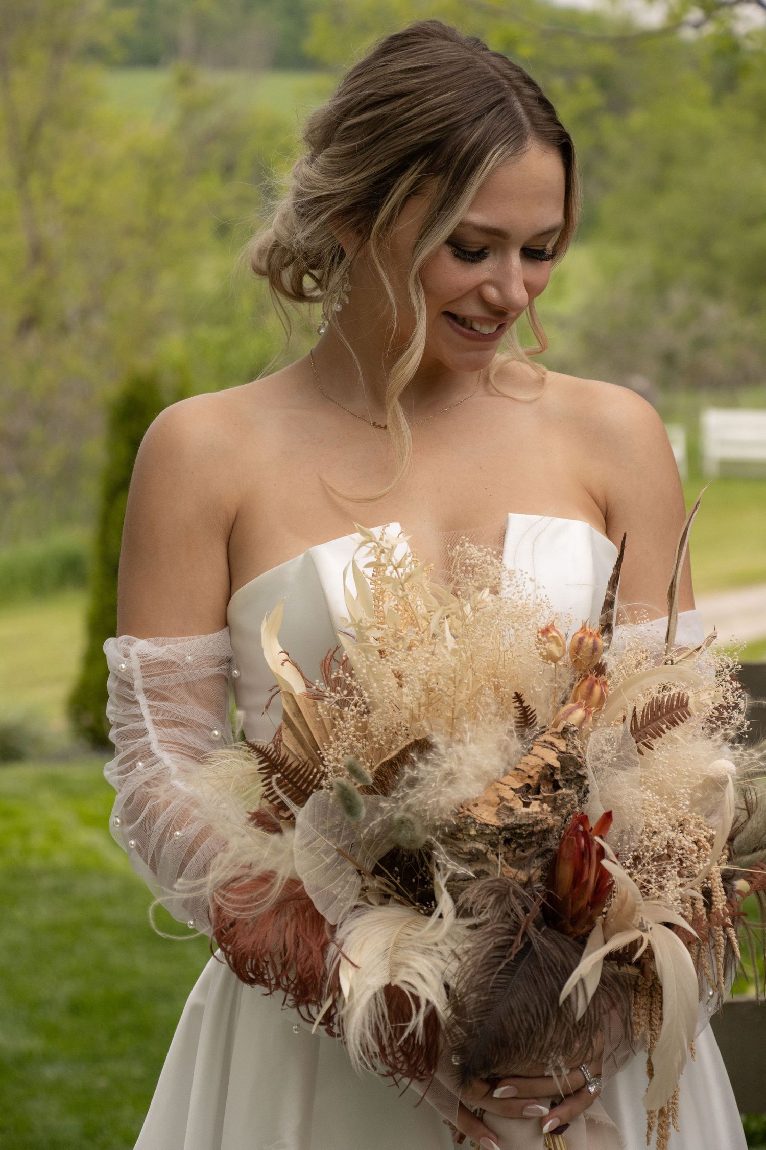 A bride wearing a strapless white gown stands outdoors, smiling softly as she looks down at an intricate bouquet. The bo