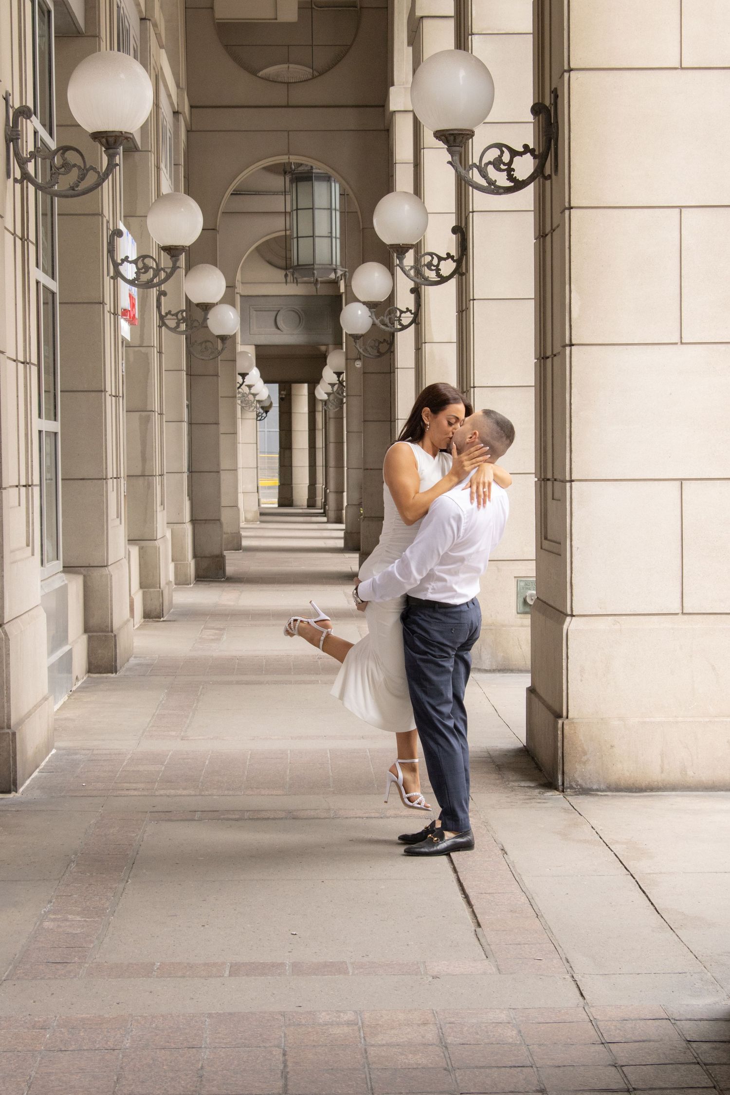 A couple shares a romantic moment on a city street. The woman, dressed in a white outfit and heels, is being lifted by