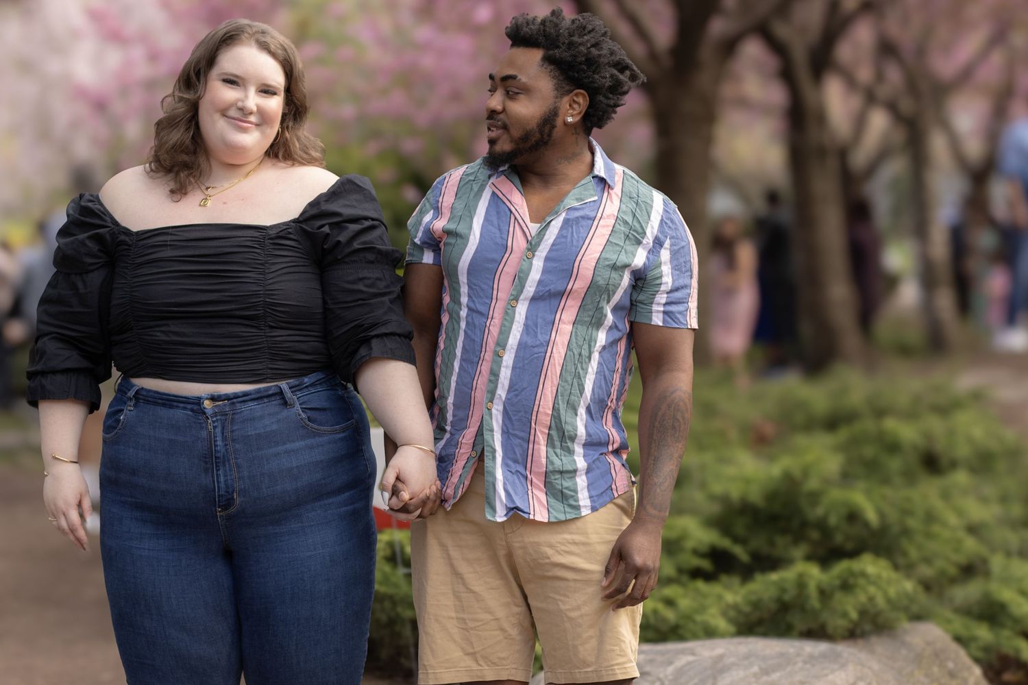 A woman in a black off-the-shoulder top and jeans holds hands with a man in a striped shirt and shorts. They are smiling