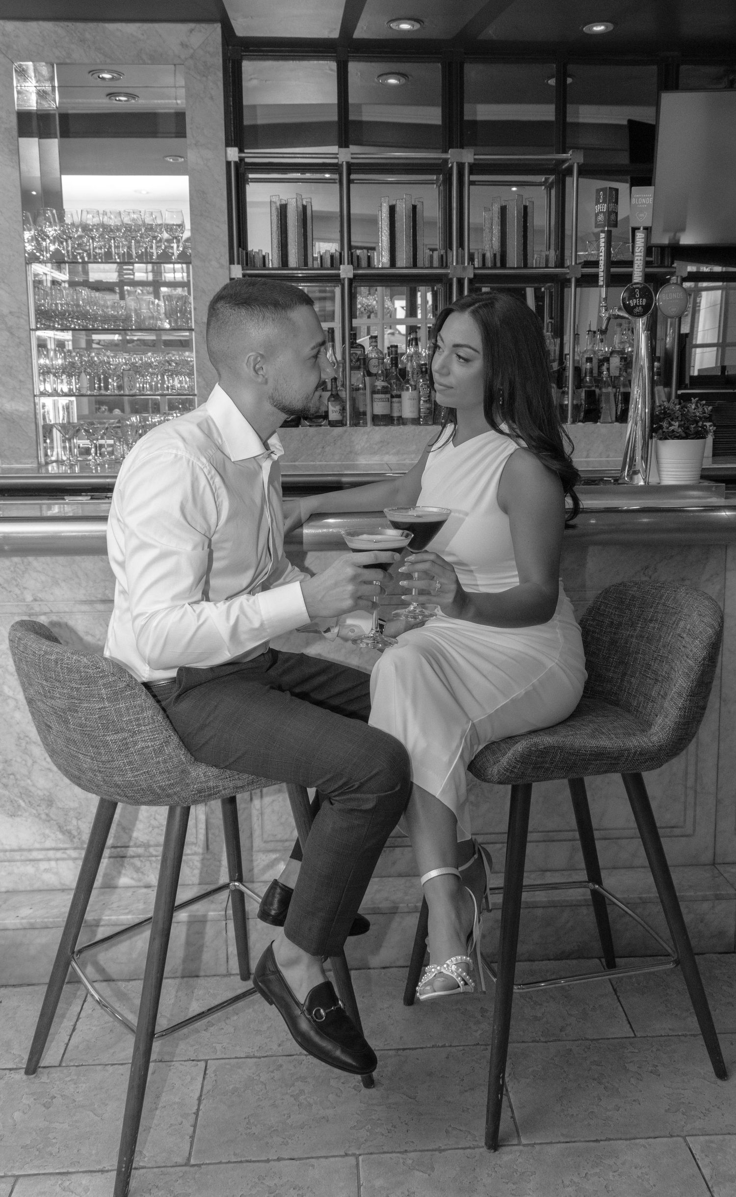 A man and a woman look at each other while sitting at a bar and holding cocktails. The man has short hair and wears a wh