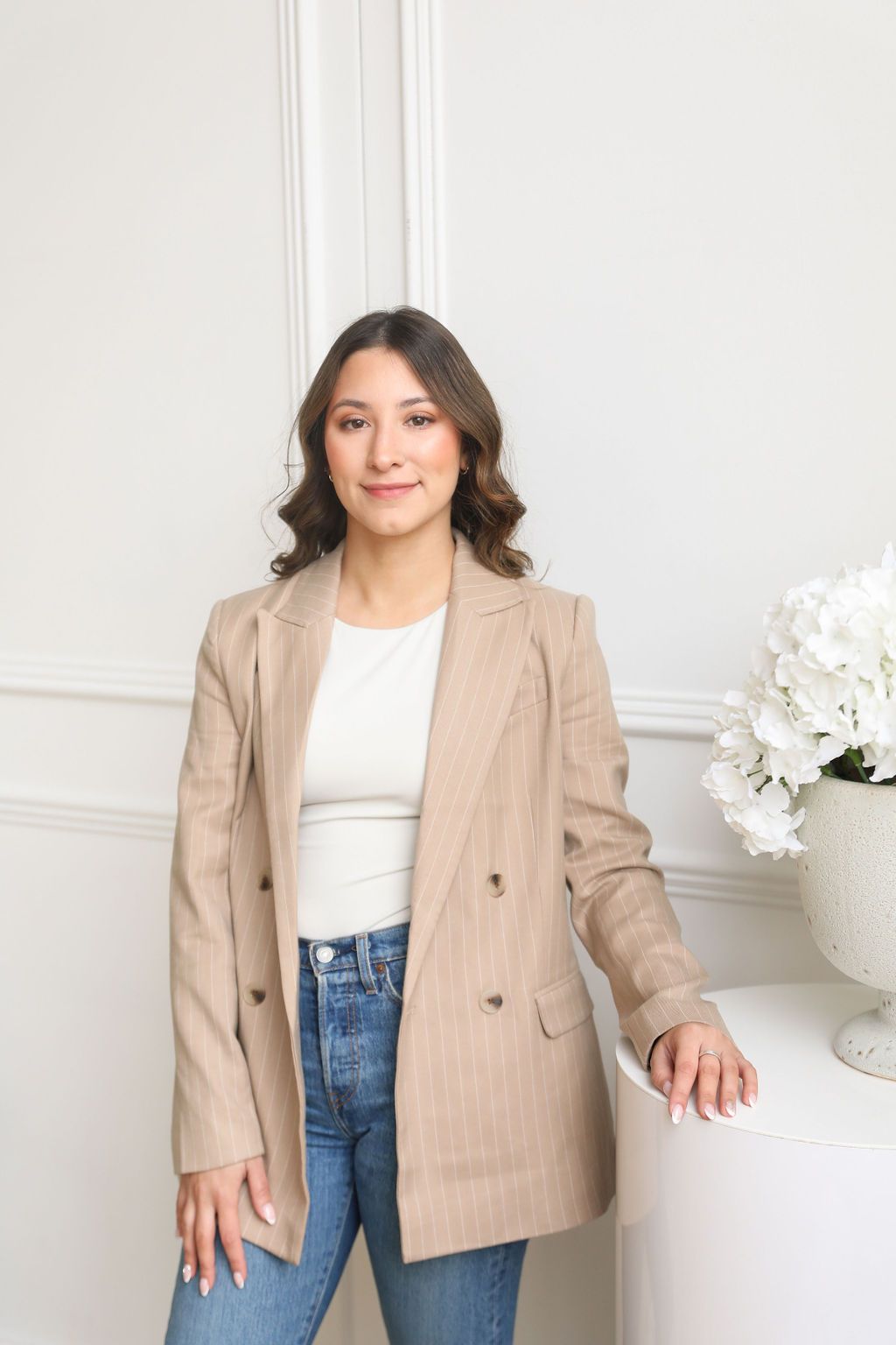 Woman in nude blazer smiling slightly, leaning on white side table with a vase of flowers.