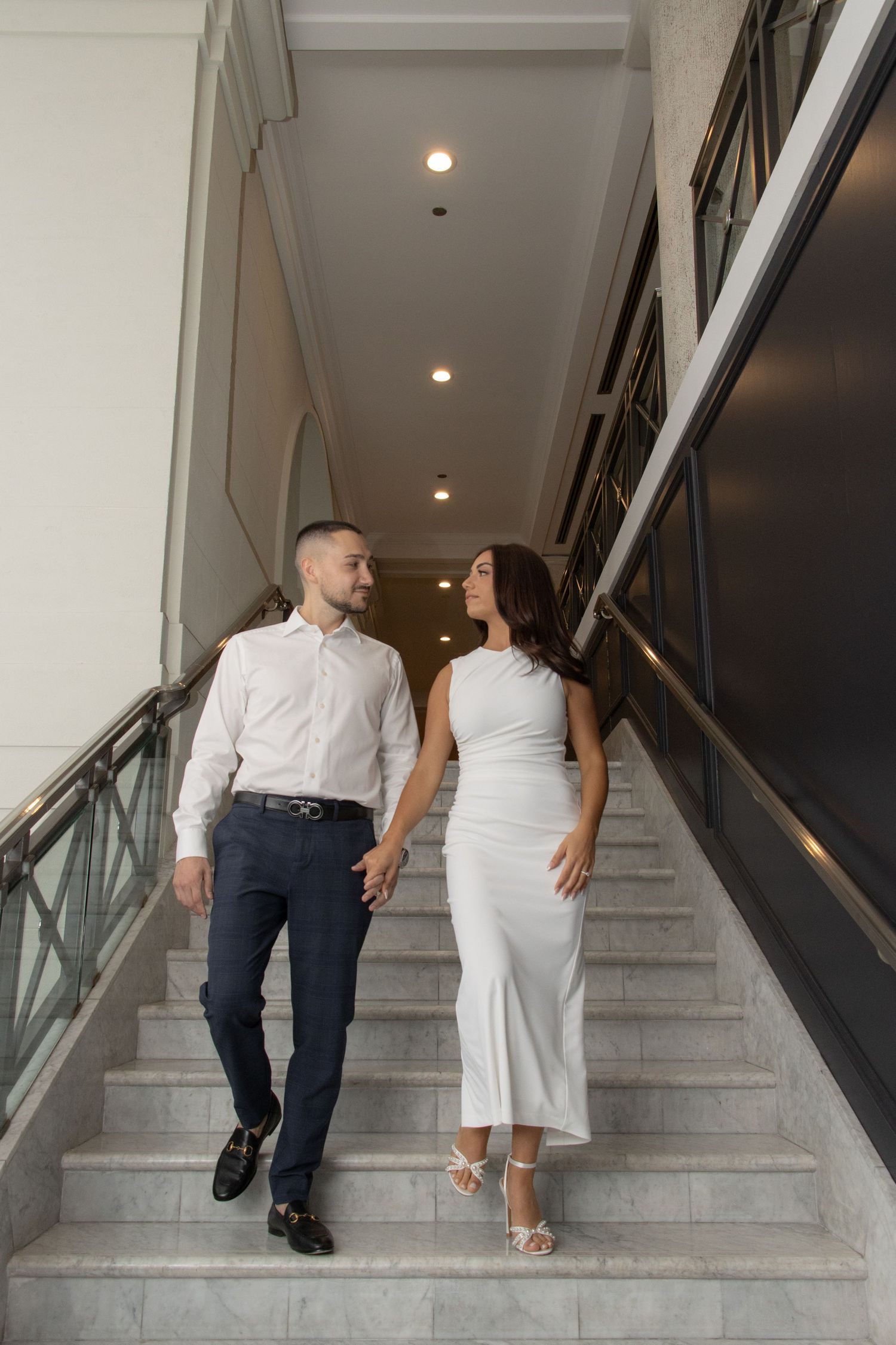 A couple holding hands while walking up a wide staircase. The woman is wearing a long, elegant dress and the man is in a